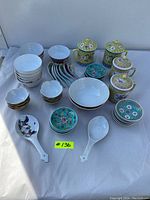 Full set of Asian porcelain serving dishes including bowls, cups with lids, and spoons arranged on a white cloth.
