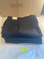 Stack of seven pairs of dark men's trousers with brand tags on top, folded and stacked on a flat surface.
