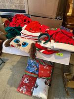 Various styles of Christmas and elf hats shown on a table, including a dog face hat and red and white themed hats. Folded flannel tablecloths with Christmas patterns are below the hats.
