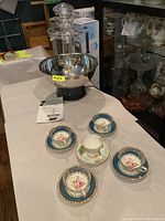 Wide view of four Royal Stafford teacups and saucers and one additional teacup with saucer along white surface next to beverage fountain.