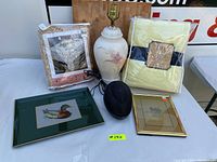 Photo showing entire lot on white surface; items include ceramic lamp, lace tablecloth, boxed crystal glass item, framed artwork, brass frame, and black speaker.