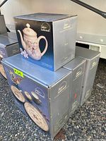 Three boxed sets of Inter-national brand stoneware including teapots and 5-piece serving dish sets stacked outdoors. Boxes are blue and gray with images of the products on the front.