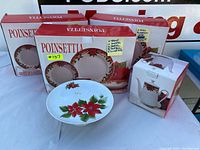 Photo showing two boxed complete Poinsettia dinnerware sets, one incomplete set, one Poinsettia cake plate, and one boxed teapot decorated with poinsettia motif.