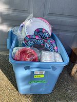 Large blue plastic bin filled with various party supplies including Happy New Year disposable party plates, red and white disposable plates, paper napkins, and plastic cups.