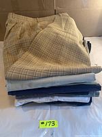 Stack of 10 pairs of men's casual pants in various patterns and colors, top pair beige and brown plaid.