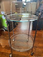 Front view of retro rolling bar cart with two glass shelves and metal frame, on wooden floor with living room background.
