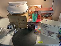 Photo showing boxed kitchen steamer, white appliance (possibly steamer parts), Dauphine colander box, green drinking glasses, metal juicer, and black circular board on a table.