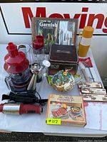 View of kitchen appliance set and related utensils including Wolfgang Puck immersion blender, measuring pitchers, lobster bibs, decorative teapot and boxed freezer containers.