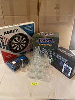 Lot displayed on table. Fourteen clear glasses arranged in a pyramid shape, boxed dartboard labeled Abbey, box labeled Hats Off (hockey socks), and boxed NHL Comfy Throw.