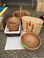 Five wicker and straw baskets and bins displayed on ground. Includes picnic basket with leather handles, cotton-lined rectangular bin, cylindrical basket, and two round baskets.
