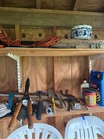 View of two wooden shelves in backyard shed showing hedge clippers, hand tools, hammer, extension cord coiled on top shelf, ceramic pot, and other items scattered on the bottom shelf.