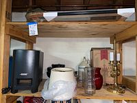 Photo of Bose Acoustic Module, various glass items, a brass candelabra-style lamp with three candle sockets and fabric lamp shade, ceramic jug, small planter, and other decor items on wooden shelf.