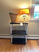 Brass tone 3-bulb lamp, black collapsible 3-tier plastic shelf, and wicker trash can arranged together on hardwood floor against a wall.