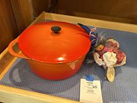Orange Cousances stock pot with lid and black knob handle, with traditional witch doll beside it on blue cloth.