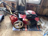 Full side view of the red Craftsman push lawnmower showing the white wheels, handle, and attached gas can next to it in a wooden shed.
