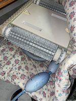 Side view of Dolphin robotic pool vacuum showing caterpillar treads and float attachments, placed on floral cushioned surface