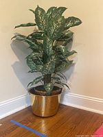Large faux plant with green and cream leaves in gold metal pot placed on wooden floor against wall.