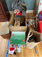 Overview of multiple boxes containing Christmas decorations, artificial wreath, outdoor lights packaging, and assorted items in bags on hardwood floor.