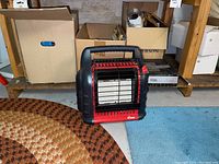 Mr Heater red and black portable space heater placed on floor near cardboard boxes and a patterned round rug.