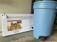 Photo showing box of freestanding pet gate labeled 'Freestanding Pet Gate Large' with gate image, and blue Rubbermaid trash can with wheels