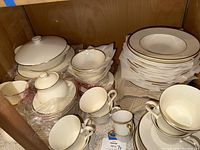 Photo showing large plates and soup bowls stacked, tea cups, cream and sugar serving pieces inside a wooden cabinet.