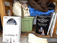 Shelf with a Sharper Image Music Responsive Massage Mat box, green storage bin, blankets, and sheets.