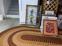 Photo showing stained glass window leaning on a braided oval rug with multiple framed art pieces around it in basement setting.