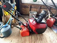 Side view of red Tornado snow thrower showing main body, handle, and one orange gas can alongside a blue watering can.
