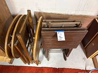 Two sets of wooden tray tables leaning against wall. One set contains three oval tables; the other set contains rectangular folding tables with dark brown tops.