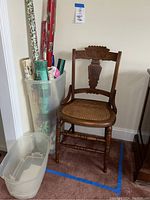 Full view of vintage wooden chair with cane seat next to a bin filled with assorted rolls of Christmas wrapping paper in various colors and patterns.