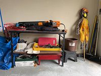 Wide shot of the lot showing the leaf blower atop a shelf, decorative scarecrow standing beside, brooms and step stool below, along with gardening tools and croquet set arranged on lower shelves.
