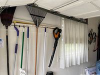 Wide view of tools hanging on garage wall including broom, rakes, broom, sweeper, hedge trimmers, blower and cords.