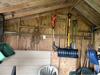 Photo of yard tools hanging on wooden shed wall showing weedwhacker, yellow snow shovel, and blue scoop shovel.