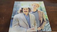 Front cover of Simon & Garfunkel Greatest Hits vinyl album showing the duo outdoors, one in tan blazer and cap, the other in striped shirt and brown jacket.