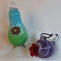 Two hummingbird feeders displayed side by side. One is purple swirled glass with red glass flower and clear handle. The other is blue-green with birdhouse motif and bronze legs.