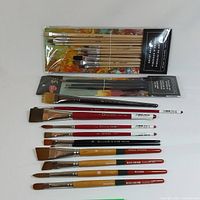 Overall layout of paint brushes including two sets in plastic packaging and multiple loose brushes of different brands and colors on white background.