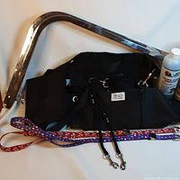 Photo showing black nylon grooming hammock with attached metal frame, grooming arms, and two colorful dog leashes with metal clips on white background.
