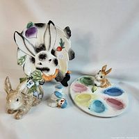 Photo showing all four Fitz and Floyd ceramic Easter-themed items together: a white bunny plate, brown rabbit figurine, small bird figurine, and egg holder tray with bunny brush.