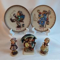 Lot of five Goebel porcelain collectibles: two annual plates displayed on white stands, three various children figurines, one with umbrella showing chips.