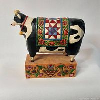 Front view of decorative cow figurine showing full body with quilt patterned blanket and floral base.