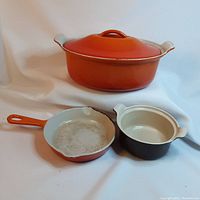 Three Le Creuset cookware pieces on white background: one covered oval pot in orange, one small orange skillet pan showing wear inside, and a small two-handled round pot in brown and cream.