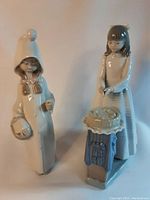 Two Lladro porcelain figurines shown together. One girl figure holding a basket; the other girl figure cutting a cake on a table with missing cutter.