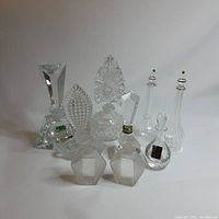 Full lot of nine empty clear glass and crystal perfume bottles with ornate stoppers, placed closely together on white background.