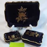 Large rectangular porcelain serving plate in cobalt blue with gold floral decoration and scalloped gilded edge shown upright on stand alongside two smaller matching trays