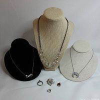 Three necklaces displayed on busts: infinity symbol on black bust, crystal bead necklace on beige bust, crescent moon pendant on beige bust, with four small rings or charms in front.