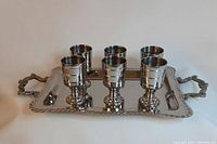 Six stainless steel shot glasses on a rectangular tray with ornate handles, showing the set from front angle.