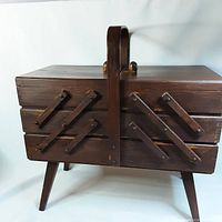 Closed view of dark wood accordion style 3-tier sewing box with dovetail corner design and single handle on top, standing on four angled legs.