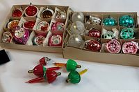 Full view of two cardboard trays containing vintage Christmas ball ornaments in assorted colors including red, gold, white, turquoise, pink. Several loose icicle-style glass light bulbs in red, green, yellow, and orange with screw bases lie in front.