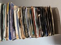 Top view of box filled with mixed 45 RPM vinyl records in paper sleeves, showing worn condition.