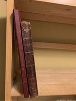 Two hardcover books standing on a wooden shelf, one dark brown and worn (ZaZa Opera 1905), one red hardcover (manuscript 1979).